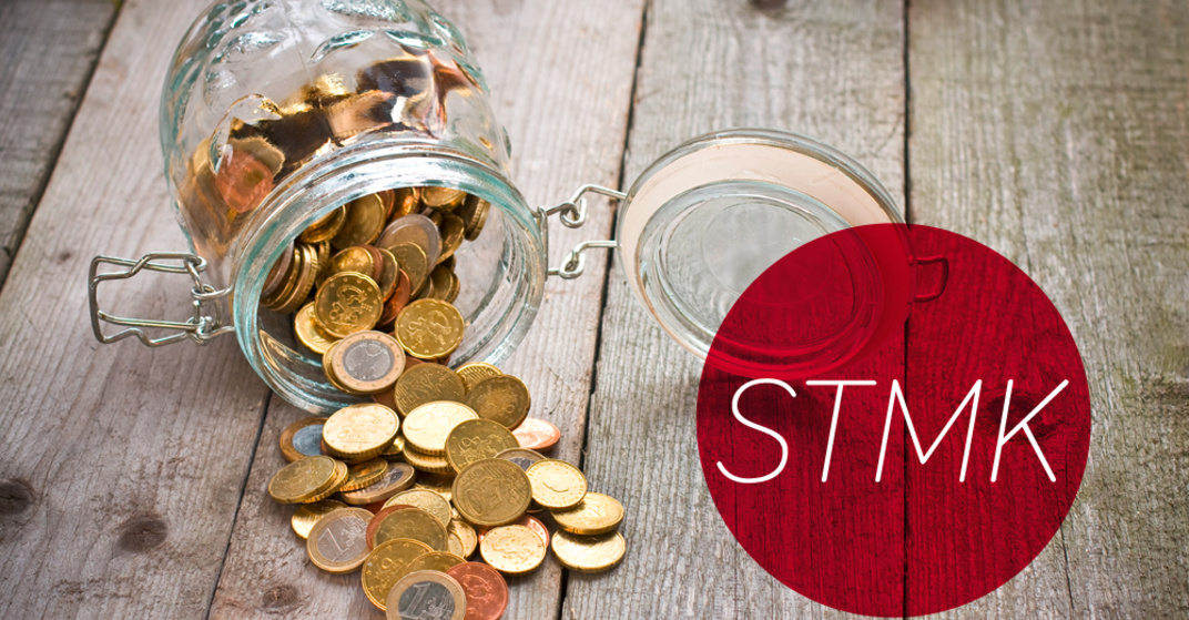 Gekipptes Einmachglas mit verschiedenen Euro- und Centmünzen liegt umgekippt auf einem rustikalen Holzboden, dazu der Text "STMK" in rotem Kreis.