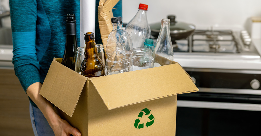 Sportlich gekleidete Frau trägt einen übervollen Recycling-Karton mit leeren Plastik- und Glasflaschen