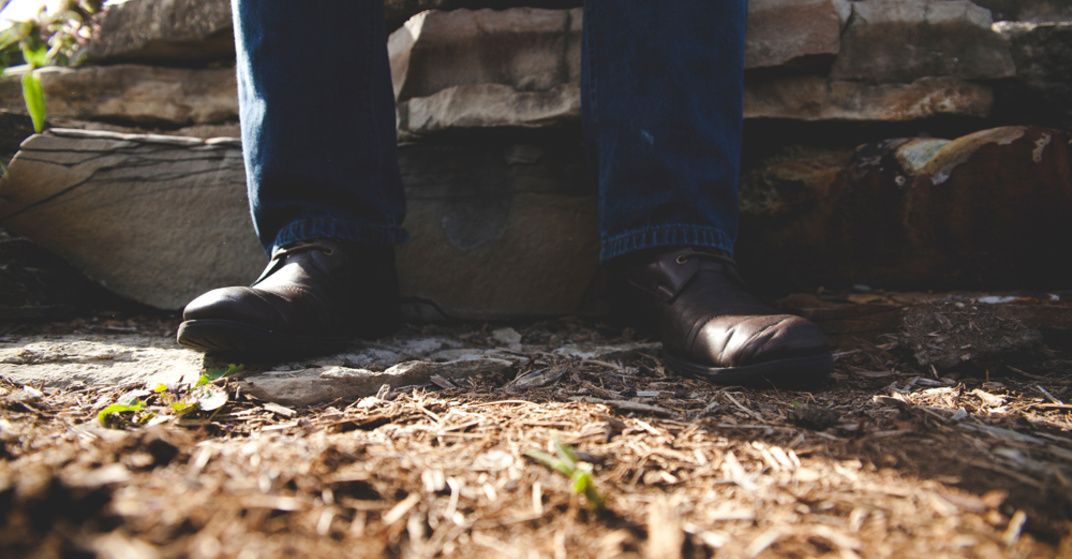Mann mit blauer Jeans und braunen, groben Lederstiefeln sitzt auf niedriger Steinmauer, nur bis Knie sichtbar, natürlicher Waldboden
